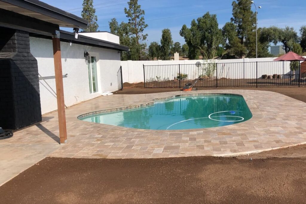 Backyard kidney-shaped swimming pool with travertine paver patio and safety fence at home, landscaping under renovation.